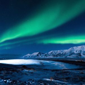 L'aurora boreale verde risplende sulle montagne innevate e su un paesaggio ghiacciato sotto un cielo notturno stellato.