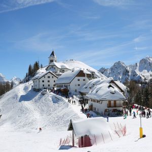 Un villaggio alpino innevato con una chiesa su una collina, circondato da sciatori e vette innevate sotto un cielo azzurro brillante.