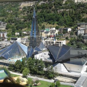 Veduta aerea di un moderno complesso architettonico con piramidi di vetro e un'alta guglia, situato in una città ai piedi di una montagna verde e boscosa.