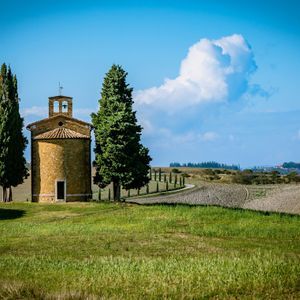 Una cappella in pietra con campanile si erge tra due cipressi su una collina verde sotto un cielo azzurro con nuvole.