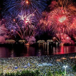 Un enorme spettacolo di fuochi d'artificio illumina il cielo notturno sopra una spiaggia affollata di persone che osservano dalla riva.
