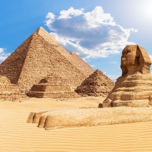La Grande Sfinge e tre grandi piramidi si ergono nel deserto sotto un cielo azzurro e soleggiato, con una persona e un cammello in primo piano.