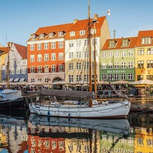 Barche a vela ormeggiate in un canale di fronte a edifici colorati sul lungomare, con i loro riflessi che brillano sull'acqua.