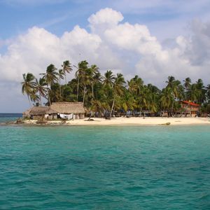 A small tropical island with palm trees and huts on a white sand beach, surrounded by turquoise water under a cloudy sky.