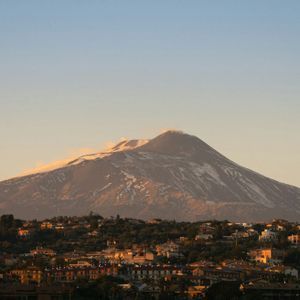 Un grande vulcano innevato, con un pennacchio di fumo dalla sua cima, si erge maestoso su una cittadina collinare bagnata dalla calda luce dell'alba.