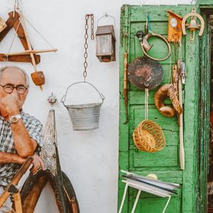 Un anziano signore siede pensieroso fuori da un negozio, la cui parete bianca e la porta verde sono decorate con vari oggetti vintage.