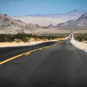 Una carretera pavimentada con líneas amarillas se extiende por un valle desértico, con montañas rocosas visibles a lo lejos bajo un cielo azul.