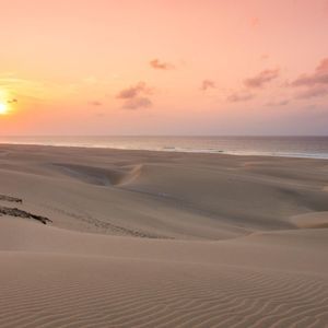 El sol se pone sobre el océano, proyectando un resplandor rosa y naranja sobre un vasto paisaje de dunas de arena.