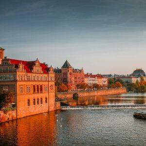 Edifici storici con tetti rossi e una torre dell'orologio costeggiano un fiume durante un tramonto dorato, con un battello turistico nell'acqua.