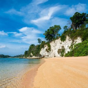 Una spiaggia sabbiosa con acqua calma e limpida accanto a una scogliera di roccia bianca sormontata da alberi verdi lussureggianti sotto un cielo blu.