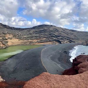 Una vista panoramica di una laguna verde accanto a una spiaggia di sabbia nera, con una montagna vulcanica stratificata e il mare sotto un cielo nuvoloso.