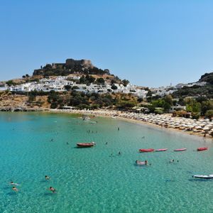 Una vista aerea di una baia con acqua turchese, una spiaggia sabbiosa con ombrelloni bianchi e un villaggio collinare con una fortezza in cima.