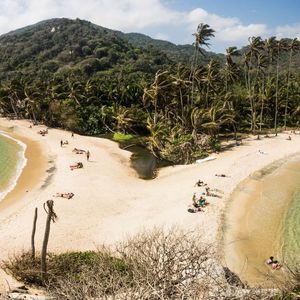 Un uomo siede su una grande roccia che domina due spiagge tropicali con acqua turchese, palme e una lussureggiante collina verde.