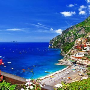 Un pueblo colorido en un acantilado verde con vistas a un mar azul salpicado de barcos, con flores brillantes en primer plano.