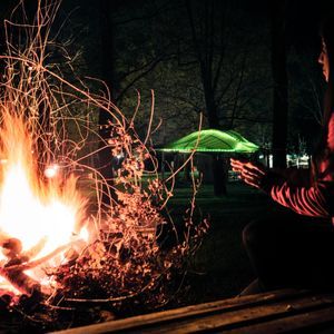 Una persona si siede a scaldarsi le mani accanto a un grande falò di notte, con scintille a lunga esposizione che creano scie luminose nel cielo scuro.