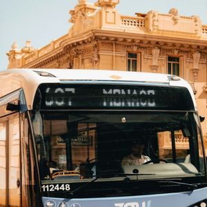 Vista frontale di un autobus blu e bianco su una strada cittadina, con un autista al volante e un edificio giallo sullo sfondo.