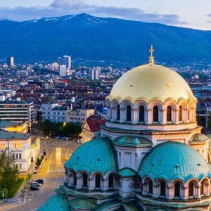 Una cattedrale dalle cupole dorate e verde acqua domina lo skyline di una città al tramonto, con un'ampia catena montuosa sullo sfondo.