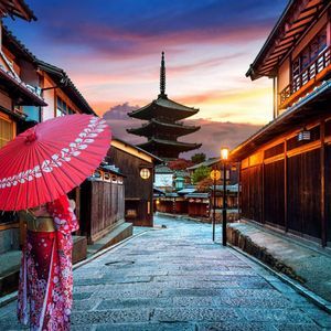 Una persona en kimono con un paraguas rojo en una calle de piedra de un pueblo tradicional, con una pagoda visible al atardecer.