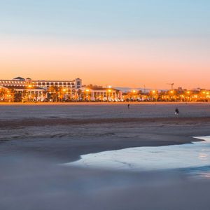 Ampia spiaggia sabbiosa al tramonto, con la battigia in primo piano e uno skyline cittadino illuminato sotto un cielo arancione e blu.