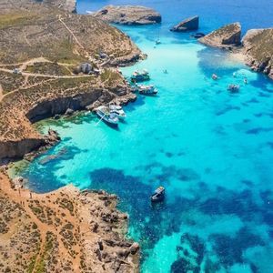 Veduta aerea di una costa rocciosa con una luminosa laguna turchese dove sono ormeggiate diverse imbarcazioni e persone nuotano.