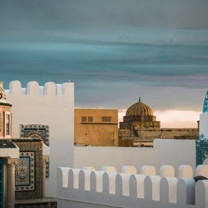 Two ornately decorated domes in blue and orange sit atop whitewashed buildings with crenellated walls under a cloudy sky.