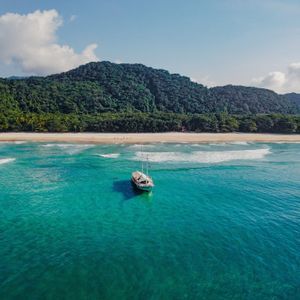 Una vista aérea de un barco blanco anclado en agua turquesa, con una playa de arena y una exuberante montaña verde al fondo.