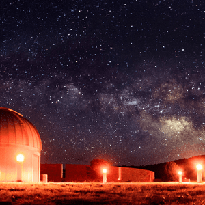 Un osservatorio astronomico, illuminato da calde luci arancioni, si trova sotto un cielo notturno sereno pieno di stelle e della Via Lattea.
