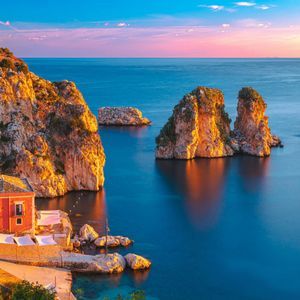 Un pueblo costero y una torre de piedra sobre acantilados soleados, con vistas a aguas azules y tranquilas y farallones al atardecer.