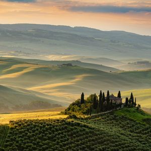 Una villa in pietra circondata da cipressi si erge su una collina che domina un paesaggio di dolci colline verdi all'alba.