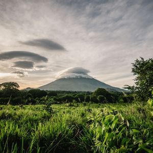 Un volcan avec un nuage lenticulaire au-dessus de son sommet, s'élevant au-dessus d'un paysage verdoyant de champs et de forêt au coucher du soleil.