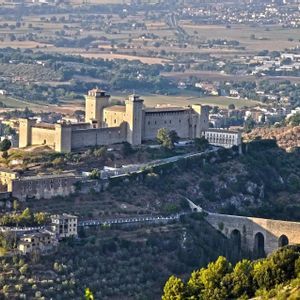 Un gran castillo de piedra con torres se asienta sobre una colina verde con vistas a un valle y un puente arqueado.