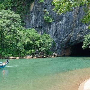 Una barca turistica blu con persone naviga su un fiume turchese, dirigendosi verso una grande apertura di grotta alla base di una montagna boscosa.