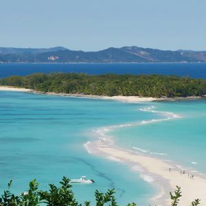 Eine sich schlängelnde weiße Sandbank verbindet sich mit einer kleinen, grünen Insel in türkisfarbenem Wasser, mit fernen Bergen im Hintergrund.