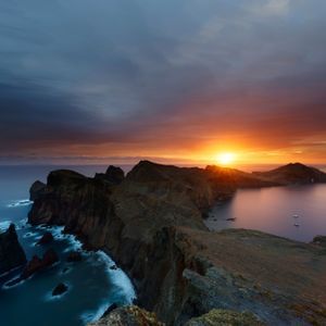 Una penisola rocciosa si estende nell'oceano sotto un cielo drammatico mentre il sole sorge sull'acqua.