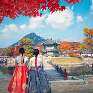 Two women in traditional dresses stand before a lake looking at a pavilion, with vibrant red autumn leaves framing the scene.