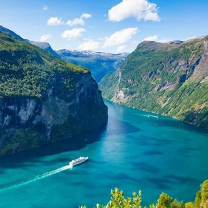 Vista aérea de un crucero blanco navegando por un fiordo turquesa, rodeado de escarpadas montañas verdes bajo un cielo azul.