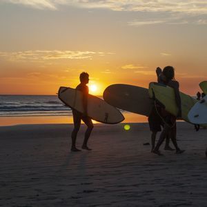 Un viaggio di gruppo WeRoad di surfisti in controluce mentre camminano lungo una spiaggia portando le loro tavole da surf al tramonto.