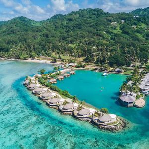 Vista aerea di bungalow sull'acqua che circondano una laguna turchese, con un'isola tropicale lussureggiante sullo sfondo.