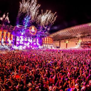 Una folla immensa con le mani alzate di fronte a un palco illuminato a giorno con fuochi d'artificio sopra al festival musicale Ultra Europe di notte.