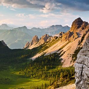 La luce del sole colpisce le cime di una catena montuosa rocciosa che si erge su una valle verde con una foresta, sotto un cielo nuvoloso.