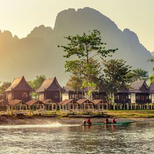 Una barca de cola larga con tres personas navega por un río pasando casas de madera sobre pilotes al atardecer, con montañas difusas al fondo.