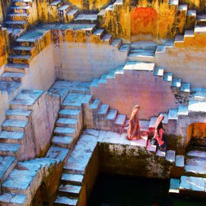 Dos mujeres con vestimenta tradicional descienden las escalinatas de piedra geométricas de un antiguo pozo escalonado.
