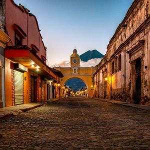 Una calle empedrada vacía al anochecer conduce a un arco amarillo con una torre del reloj, con un volcán visible a lo lejos.