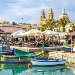 Des bateaux colorés sont amarrés dans un port, à côté de cafés en bord de mer, avec une église et la ville en arrière-plan.