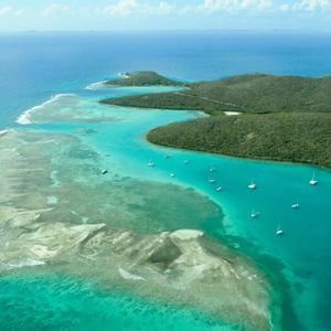 Una vista aerea di barche a vela ancorate in una baia turchese accanto a un'isola verde con una barriera corallina visibile.
