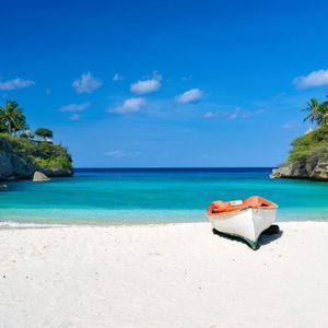 Una piccola barca si trova su una spiaggia di sabbia bianca in una caletta con acqua turchese e scogliere sovrastate da palme.