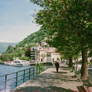 Una persona passeggia lungo un viale alberato e ombreggiato, accanto a un lago con barche ormeggiate e una cittadina collinare.