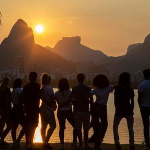 A WeRoad group trip stands silhouetted on a lakeshore, watching the sunset over mountains and a city.