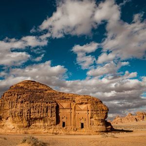 Un'antica tomba scavata in un grande monolite di arenaria nel deserto sotto un cielo azzurro brillante con nuvole bianche.