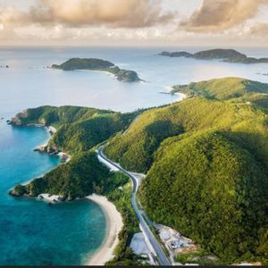 Una vista aérea de una carretera sinuosa a lo largo de una costa exuberante y verde con agua turquesa, calas de arena e islas en la distancia.